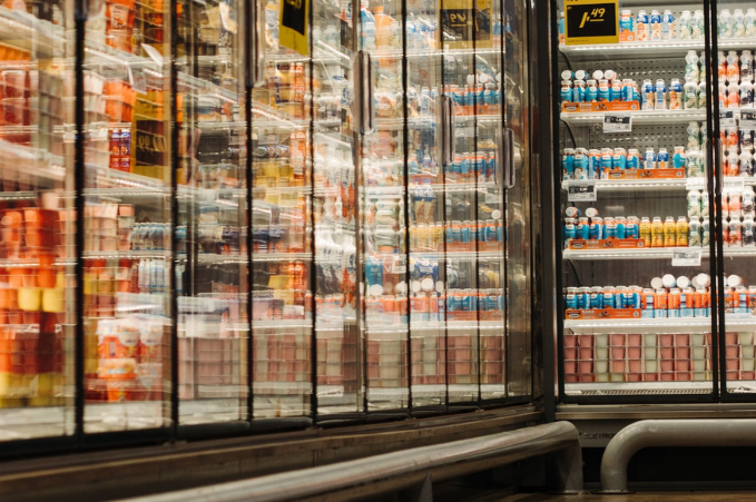 Supermarket refrigerated section with glass doors showing neatly arranged beverages including water, sports drinks, and juices, with visible price tags.