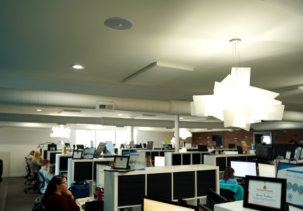 An open-plan office space with individuals working at cubicle desks, modern lighting fixtures above, and exposed brick walls, creating a contemporary industrial atmosphere.