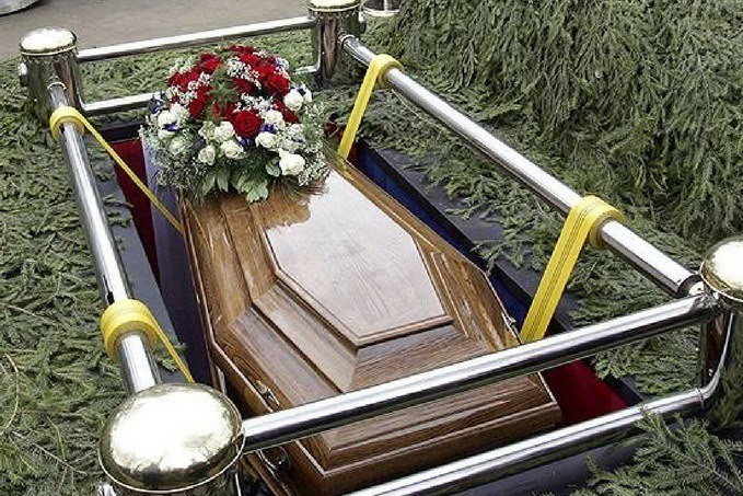 A polished wooden coffin with a floral arrangement on top, secured within a metal framework with yellow straps, set against a greenery backdrop.