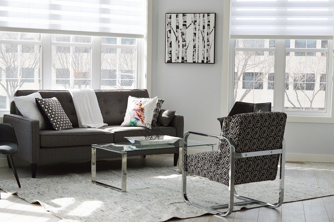 A modern living room with a dark gray sofa, patterned armchair, glass coffee table, white area rug, and a black and white abstract painting on the wall, illuminated by natural light from large windows.