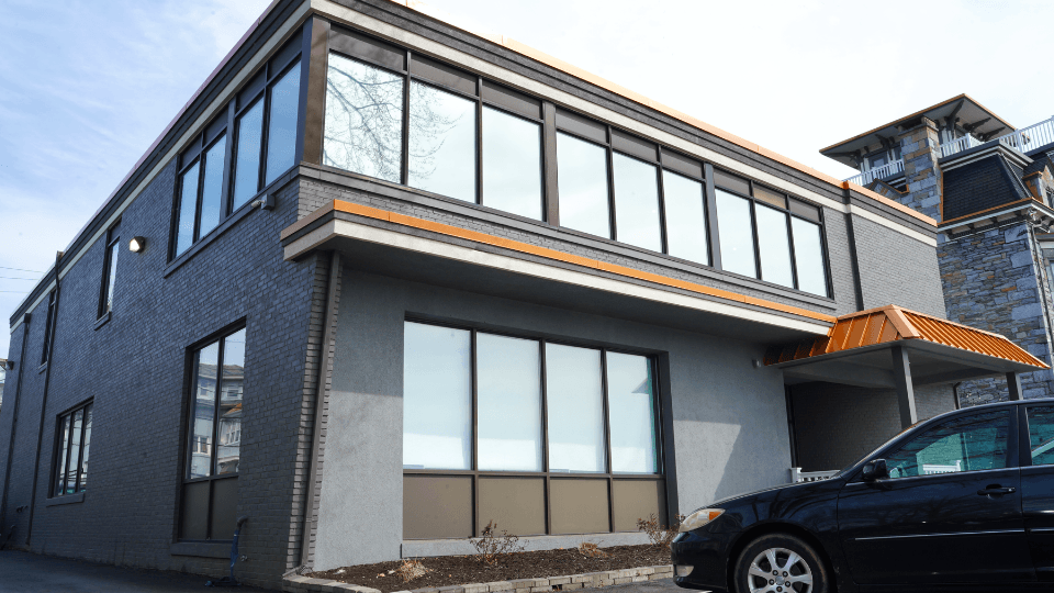 Modern two-story building with gray brick exterior, large windows, wood trim, and an orange awning over the entrance, with a black car parked in front.