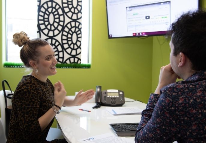 A woman and a man seated at a desk in a brightly colored office, with the woman gesturing while talking and the man listening. Office items and a computer monitor are visible on the desk.