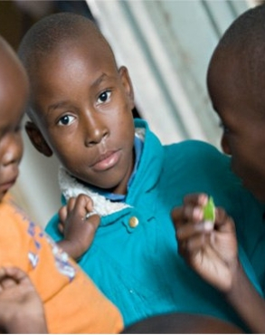 A thoughtful young child in a teal shirt looks directly at the camera, with two other partially visible children interacting nearby.