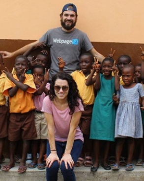 Two adults posing with a group of school children in uniforms, who are waving and making peace signs, all smiling in front of a building wall.