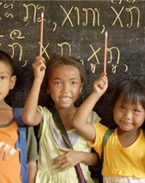 Three children in a classroom, with the middle child pointing at a chalkboard with written characters, flanked by two other smiling children.