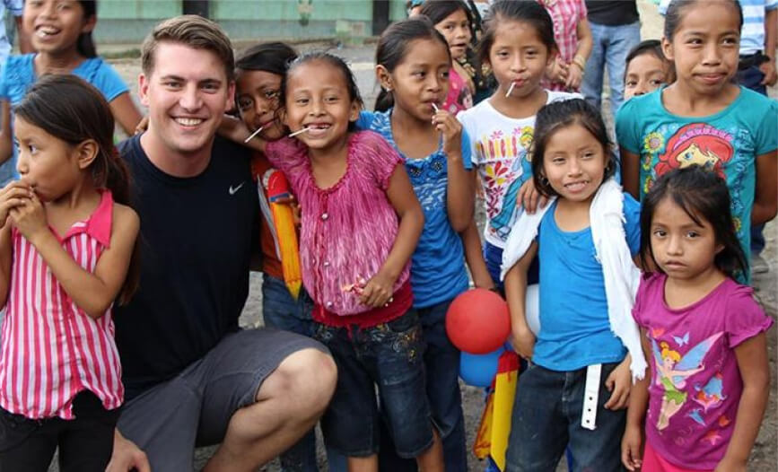 A smiling young man in a dark t-shirt crouches among a group of cheerful children of various ages, some holding lollipops, in an outdoor setting.