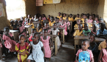 A classroom of children in school uniforms seated at wooden desks in a room with mud walls adorned with educational posters.