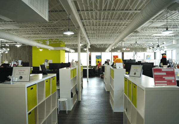 Modern open-plan office space with white desks, lime green partitions, computers, and a few people working. The space is well-lit with an industrial ceiling and colorful wall art.