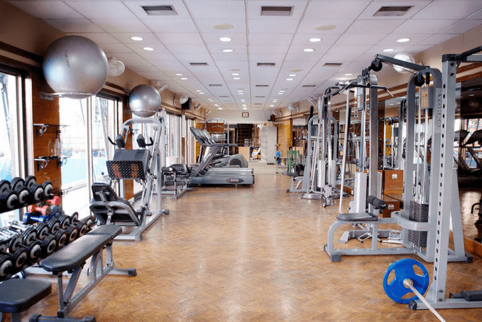 An empty gym with various exercise equipment including dumbbells, stability balls, weight machines, and treadmills, with wooden flooring and natural light.