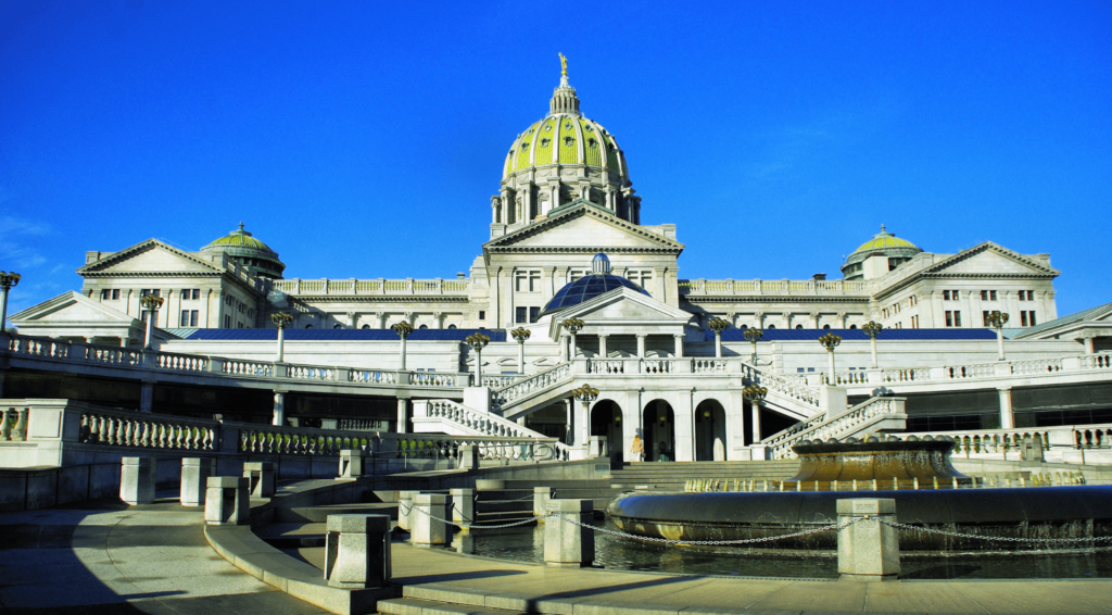 The Pennsylvania State Capitol building with a central dome, classical architecture, a fountain in the foreground, and a clear blue sky.