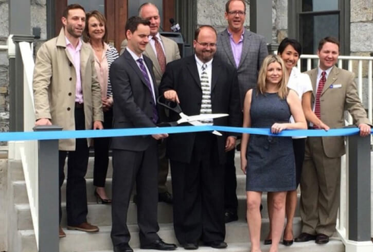 A group of nine people at a ribbon-cutting ceremony in front of a building, with two men in the center holding a large pair of scissors ready to cut a blue ribbon.