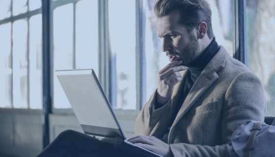 Contemplative bearded man in a coat looking at a laptop screen indoors with large windows in the background.