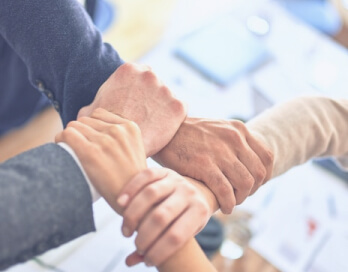 Three people's hands clasped together in a gesture of teamwork, over a blurred office background.