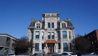 A historic three-story stone building with a mansard roof and central dormer under a clear blue sky, flanked by modern buildings, with parked cars in front.