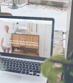 A laptop on a desk displaying a website featuring an interior designer named Brady Tolbert, with a plant in the foreground slightly out of focus.