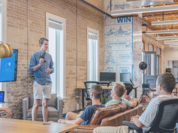 A man presenting to a small group in a contemporary office setting with exposed brick walls and motivational posters.