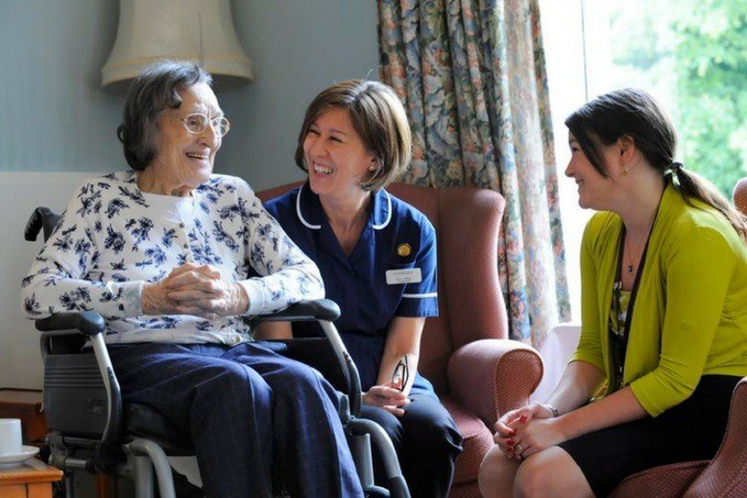 An elderly woman in a wheelchair shares a joyful moment with a laughing nurse in blue uniform and a smiling young woman in a green cardigan in a well-lit room.