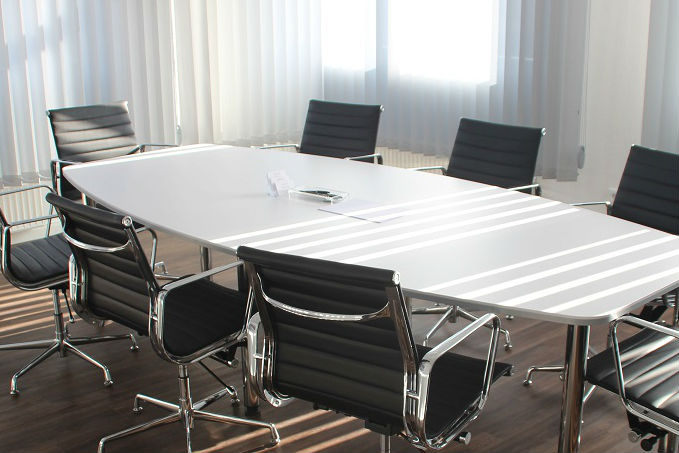 A bright conference room with a long white table, black office chairs, large windows with white curtains, and some papers and a pen on the table.