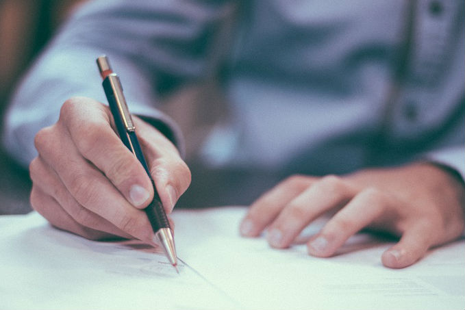 Close-up of a person's right hand writing on a piece of paper with a pen, wearing a patterned long-sleeved shirt.