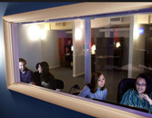 Four people in a well-lit room seen through a large window, with two conversing on the left and two looking towards the viewer on the right.