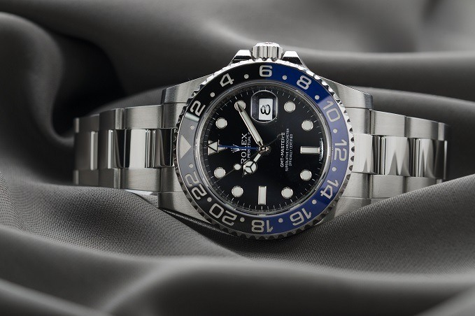 A luxury wristwatch with a stainless steel bracelet, black dial, blue and black bezel, and date function, displayed on a wavy grey fabric.