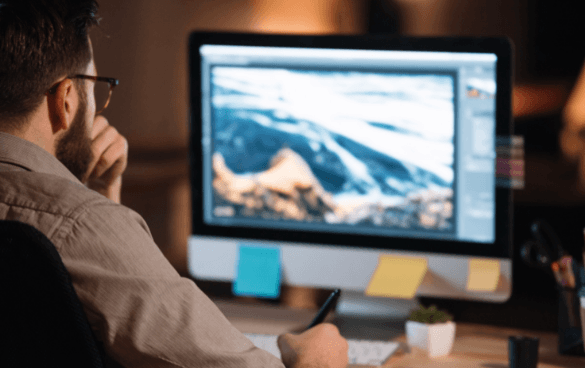 Person working on a digital drawing tablet with a graphic editing program open on the computer monitor, displaying a mountain landscape.