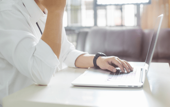 Person in a white shirt with a watch on their left wrist using a modern laptop at a desk in a softly lit room.