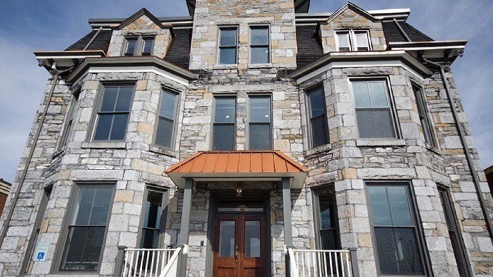 A two-story house with a stone facade, steeply pitched roof with dormer windows, symmetrical bay windows, a central wooden door with a transom window and an orange awning, white trim, and a small front porch, under a blue sky with scattered clouds.