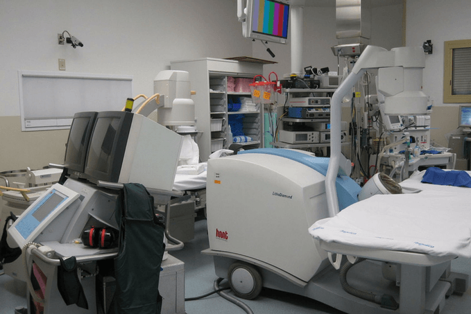 A medical operating or procedure room filled with equipment, including an imaging device, a bed, monitors, and shelves with supplies.