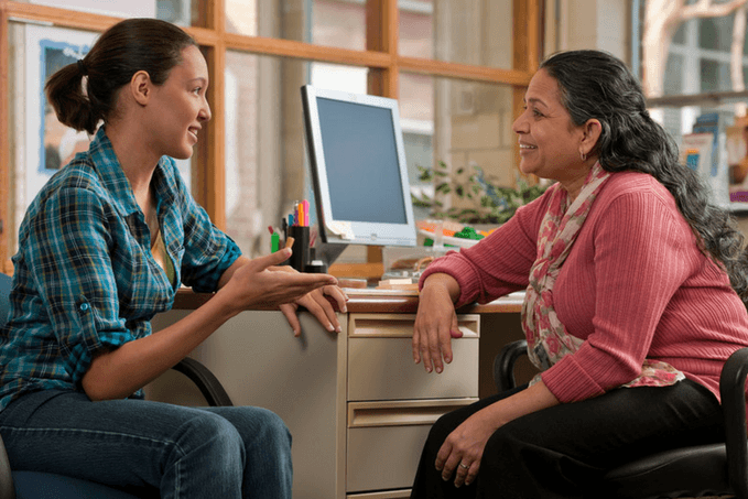 Two women are engaged in a conversation while sitting across from each other at a desk. One woman is gesturing with her hand as she speaks, and the other is listening attentively. They are in an office environment with a computer monitor, pens, and papers on the desk.