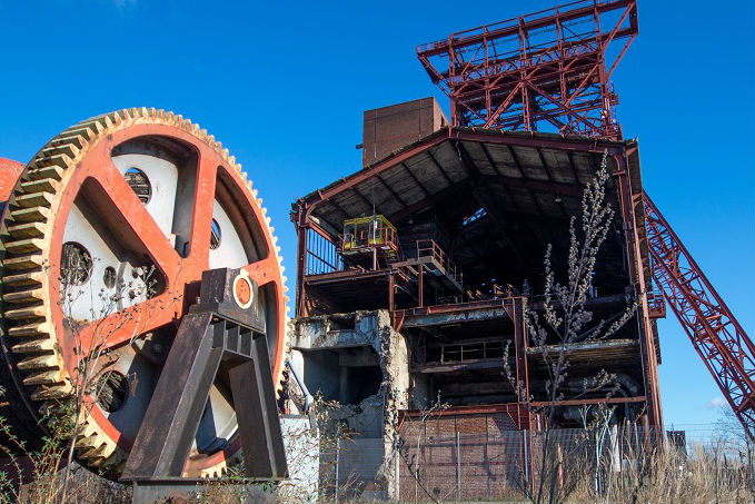 A large, rusted gear mechanism in the foreground of an abandoned industrial site with a dilapidated structure featuring a red metal framework in the background under a clear blue sky.