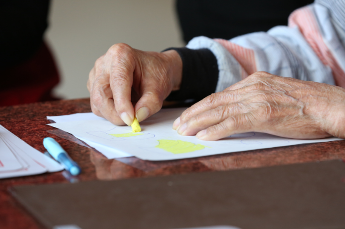 Elderly hands coloring a simple drawing with a yellow crayon, with a blue pen lying beside the paper on the table.