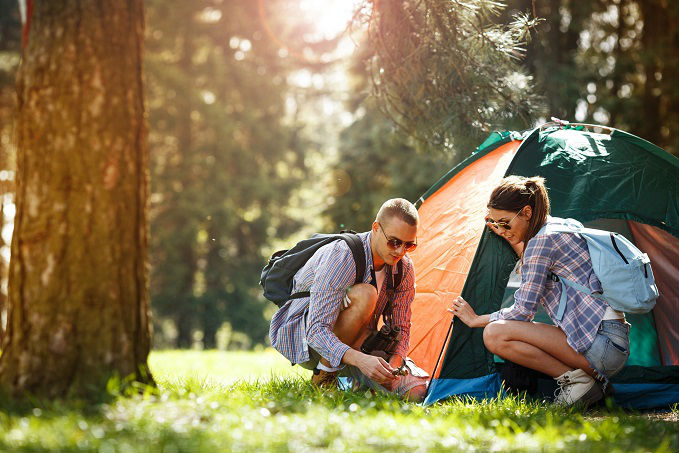 Two people setting up a tent in a sunny forest clearing, both wearing plaid shirts and backpacks.