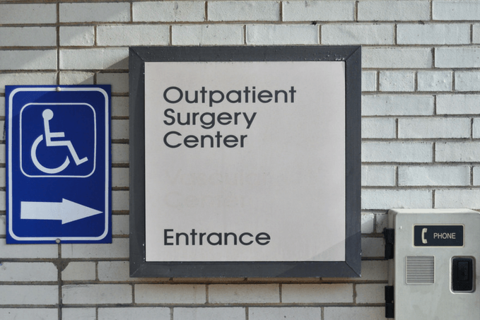 Sign on a brick wall indicating 'Outpatient Surgery Center Entrance' with an arrow pointing down, next to a handicap accessible symbol and a public telephone box.