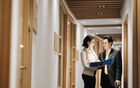 Two professionals, a man and a woman, in business attire walking through an office corridor, smiling and discussing the contents of a blue folder.