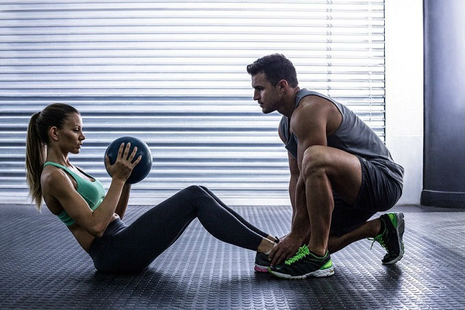 A man and a woman exercising with a medicine ball in a gym, with the man squatting and the woman sitting on the floor ready to pass the ball.