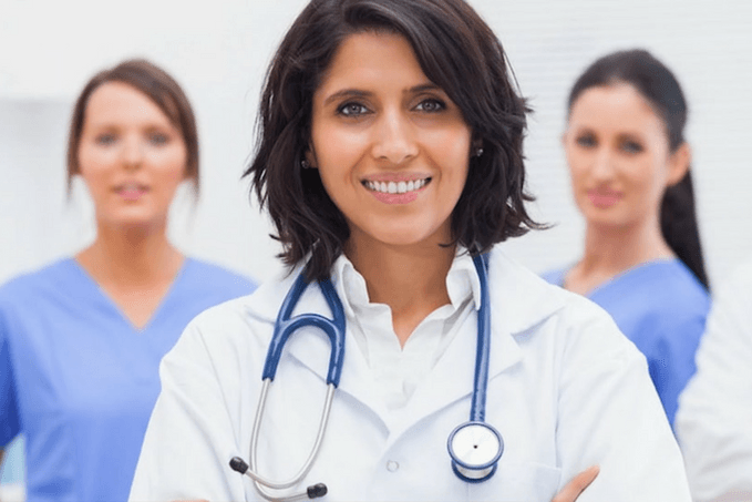 A smiling woman in a white lab coat with a stethoscope around her neck, likely a healthcare professional, with two other women in blue scrubs in the background.