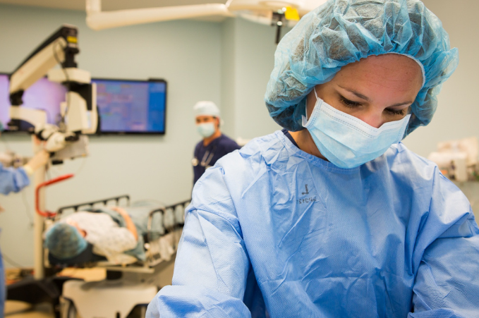 A healthcare professional in surgical attire is focused on a task in an operating room, with another medical staff member and a patient in the background.