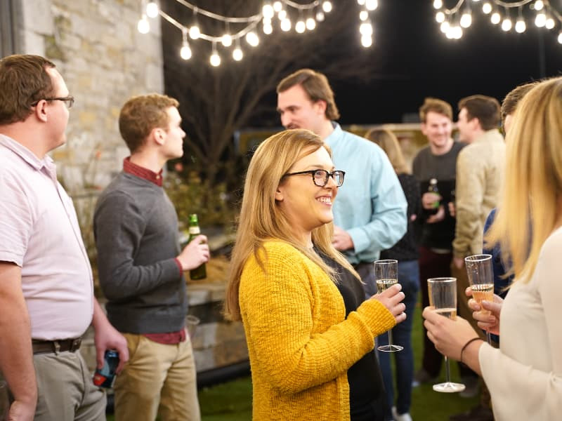 A group of people socializing at an outdoor evening event with string lights overhead, focusing on a smiling woman in a yellow sweater holding a champagne flute.