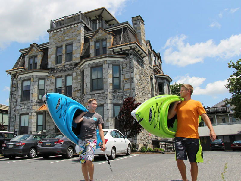 Two men carrying blue and green kayaks across a parking lot with a historic stone building in the background on a sunny day.