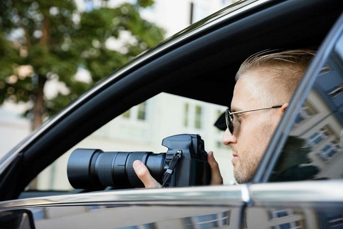 Man in sunglasses sitting in a car, looking through an open window while holding a professional camera with a long lens.