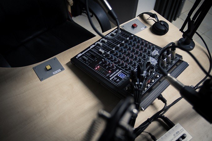 A podcasting or radio broadcasting studio setup with an audio mixing console, two microphone arms with microphones, a sound effect pad with a yellow button, and a black office chair in the background.