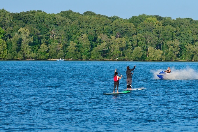 Two people paddleboarding on a lake with one person riding a personal watercraft to the right, and a forested shoreline in the background.