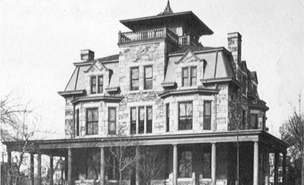 Historic stone mansion with multiple levels and a tower, featuring a wrap-around porch and surrounded by bare trees.