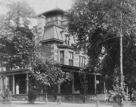 Historical black and white photo of a two-story Victorian house with a tower and wrap-around porch, surrounded by trees, with a person standing on the porch.