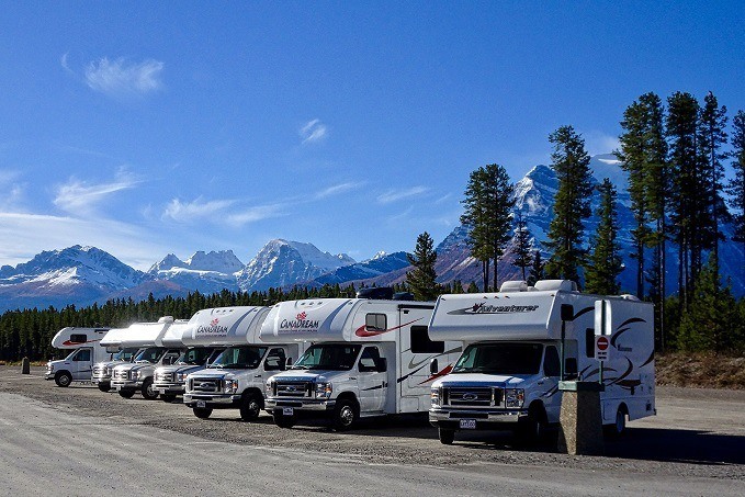 A row of 'Canadream' RVs parked on a gravel lot with snow-capped mountains and pine trees in the background under a clear blue sky.