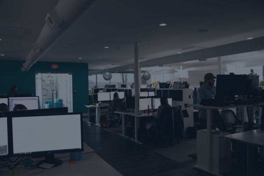 Dimly lit open-plan office space with employees working at desks with computer monitors, featuring a blue accent wall.