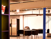 Modern office space with a desk and chair in the center, a computer monitor on the desk, a tall refrigerator to the left, and partial view of another desk to the right. The room is illuminated by natural and artificial light, with warm-toned walls and a blue column.