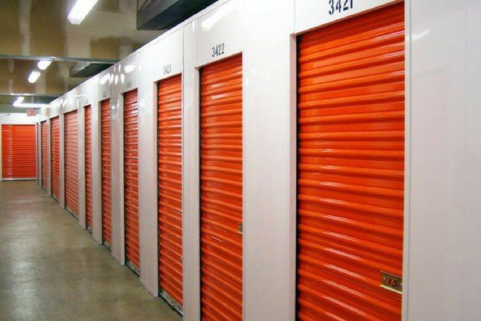 A corridor with closed red roll-up doors of storage units, numbered above each door, with white walls and bright lighting.