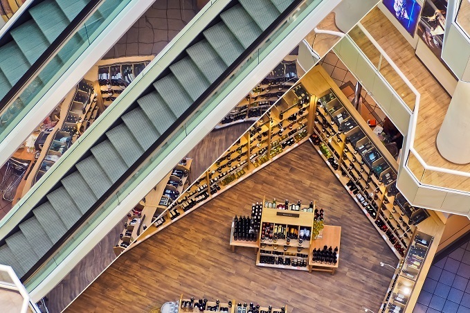 Aerial view of a shopping mall interior with two sets of escalators crossing over a wooden floor with organized retail displays of shoes and accessories.